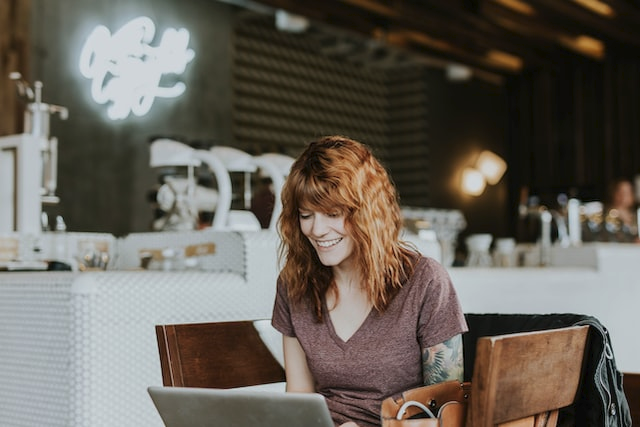 Das Bild zeigt eine lachende, rothaarige Frau am Laptop bei der Arbeit
