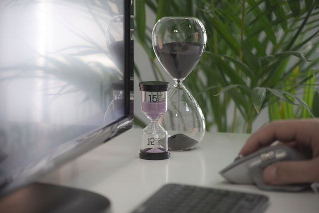 Das Bild zeigt einen Schreibtisch mit einer Sanduhr und die Hand einer Person welche einen Computer bedient